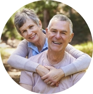 Wife with her arms around her husband who are both smiling with trees behind them